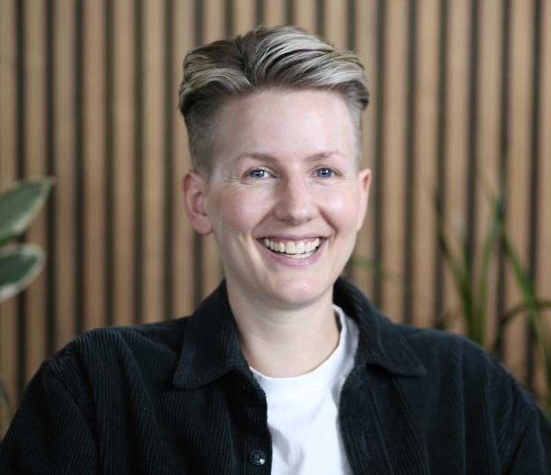 Chloe Ward-Smith is seated in front of a vertical striped background with plants in the background. They have a stort hairstyle and they are wearing a dark collared shift with a white t-shirt underneath. They are smiling and looking towards the camera.