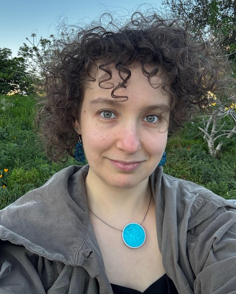 Clara Kliman-Silver who has short brown curly hair, a grey top and an eye catching turquoise necklace, she is outside in a place with lots of green plants and trees.
