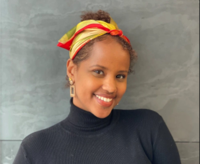 Arda Awais is smiling at the camera. She has a colourful headscarf and gold dangly earrings. She is wearing a black polo neck jumper and is front of a neutral slate-coloured background.