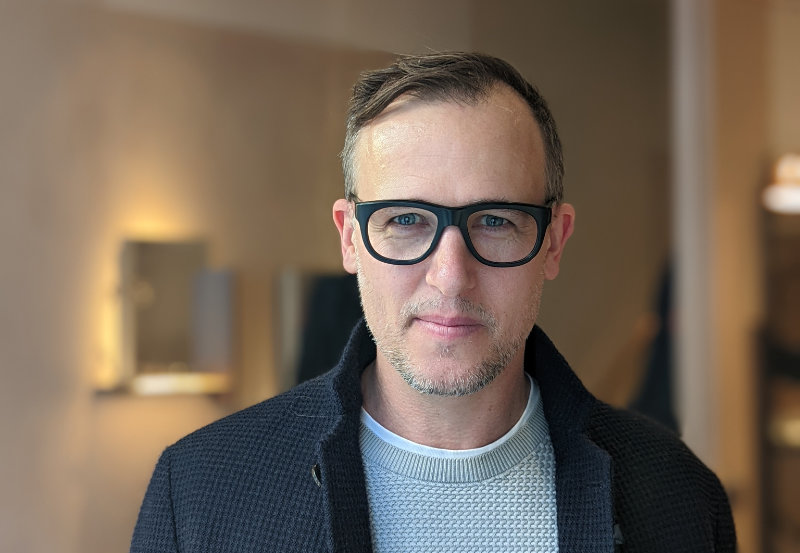 Daniel Burka is has short hair and black rimmmed glasses. He is shown in an indoor space with a blurred background. He is wearing a textured blazer and a grey woolen jumper.