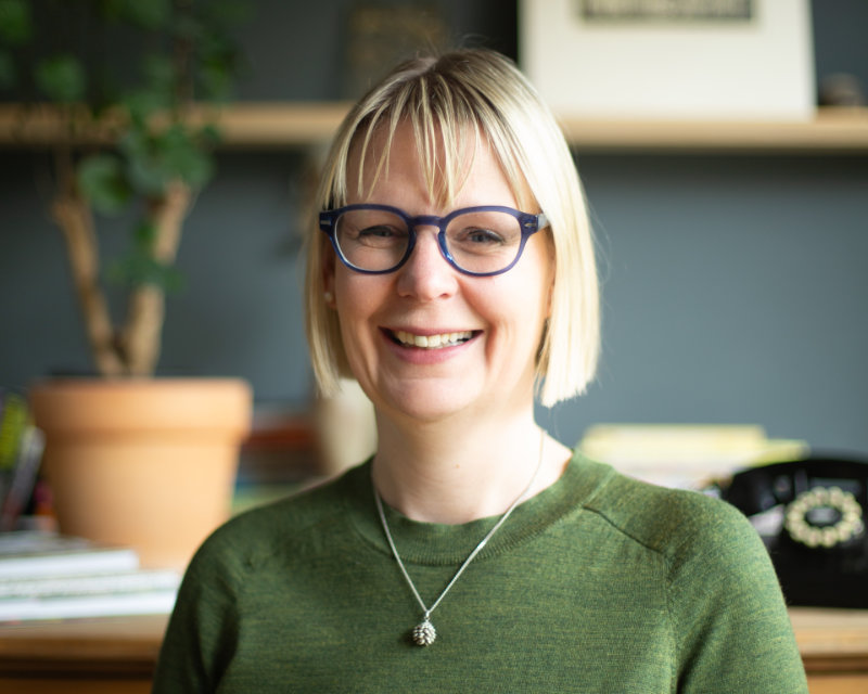Ellen de Vries is shown in an office with a plant and a rotary telephone. She has short blonde hair in a bob with a fringe. She has black rimmed glasses and is smiling. She wears a green jumper with a silver necklace.