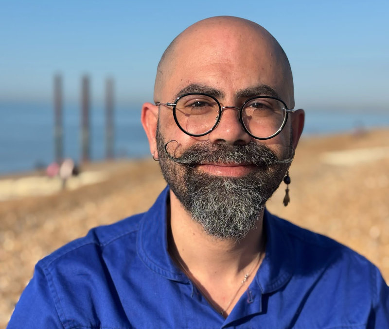 Ghaith Nassar is pictured on Brighton beach near the former West Pier. They are wearing glasses, have a styled beard and are wearing an electric blue shirt.