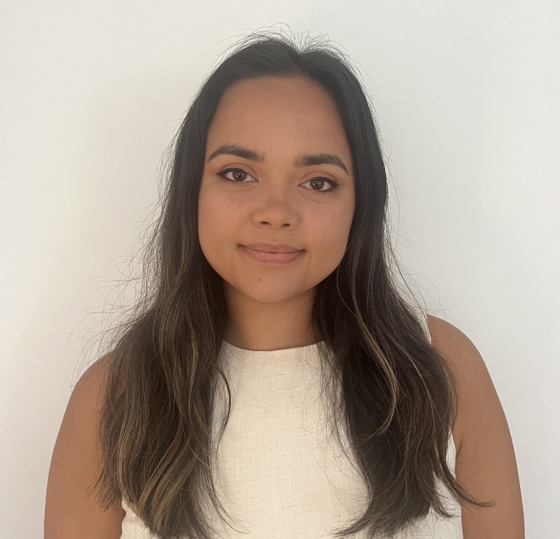 Lilymae Prescott has long brown hair and is wearing a professional white dress. She stands in front of a neutral background.