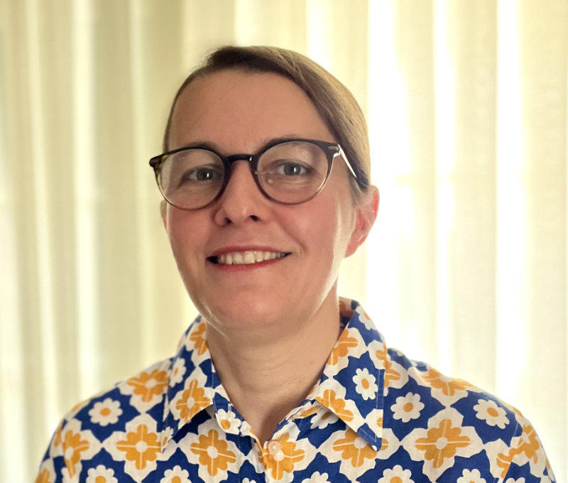 Lucy Blackwell has swept back brown hair, brown rimmed glasses and is smiling. She is wearing a brightly patterned collared shirt. There is cream-coloured curtain in the background of the image.