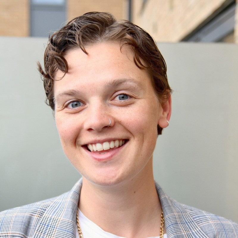 Marley Dizney Swanson has short hair and is wearing a patterns blazer. They are pictured in front of a neutral background with a building shown through a window in the background.