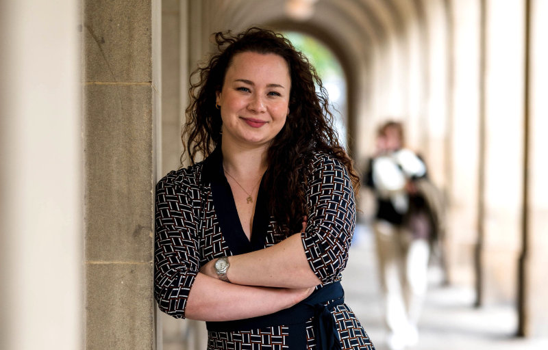 Rebecca Gordon-Watts is shown standing in a long outdoor corridor like a university campus. She is wearing a patterned dress, has curly hair and is smiling at the camera. Her arms are crossed.
