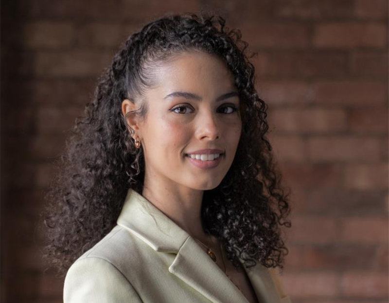 Rhea Ebanks-Simpson is shown in white blazer with curly hair swept back. She is looking directly at the camera and smiling, the background is trendy brickwork.