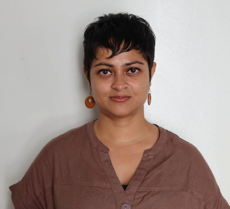 Sayani Mitra stands in front of a neutral background. She has short black hair, orange dangly earrings, and is wearing a brown shirt with big buttons.