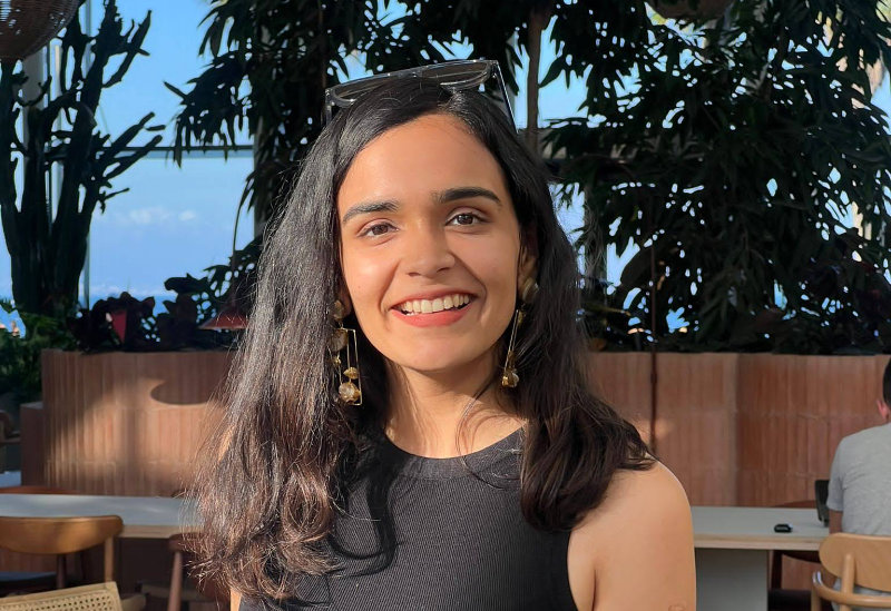 Shivani Sundararaman is outdoors in a sunny location with leafy trees in the background. She has long brown wavy hair and had a pair of sunglasses holder her hair back. She is wearing dangly earrings and is smiling at the camera.