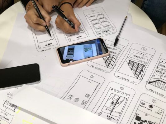 Two people are drawing wireframes based on looking at a picture on a smartphone screen. There are lots of drawn wireframes on white paper on the table already.