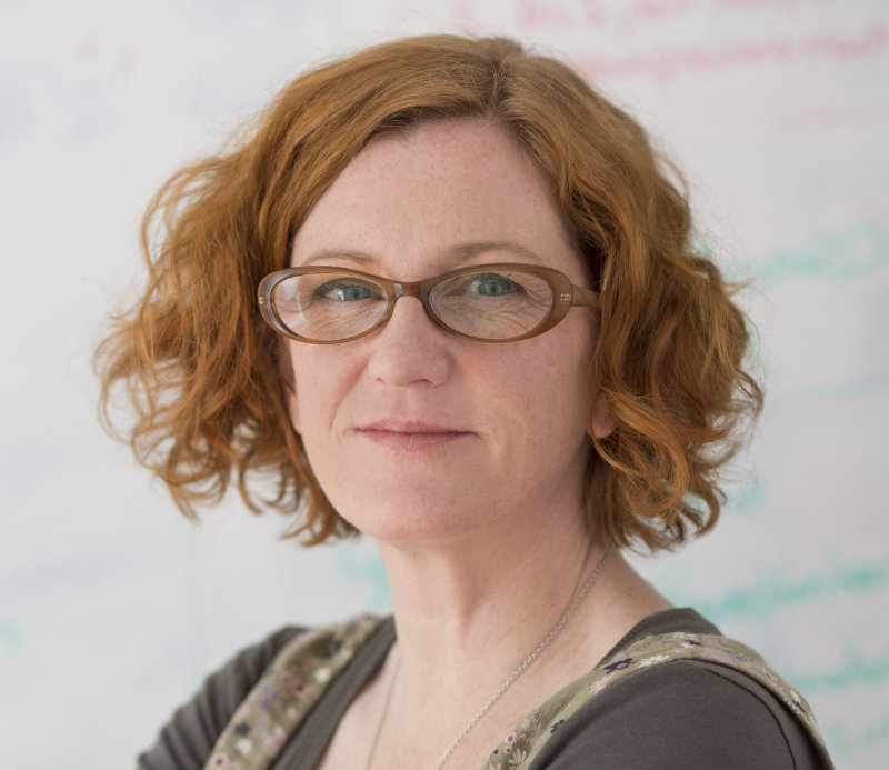Jenni Lloyd has bobbed red wavy hair, brown rimmed glasses and is wearing a necklace. She is wearing a brown top overlaid with a dress. She is standing in front of a whiteboard.