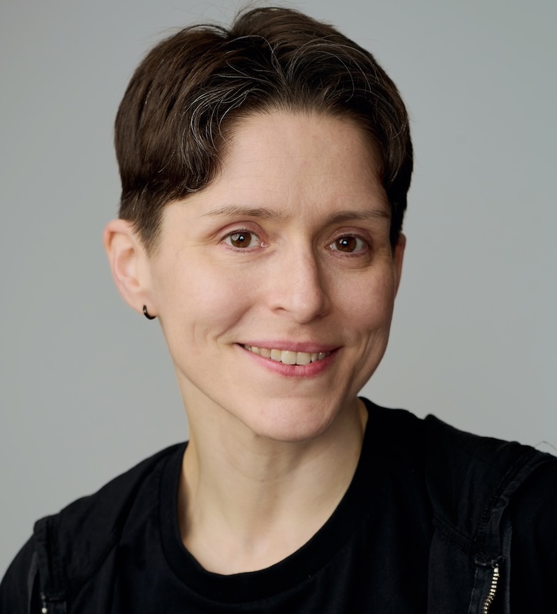 Fiona MacNeill has short hair and is wearing a black t-shirt and a black hoody and is smiling at the camera.