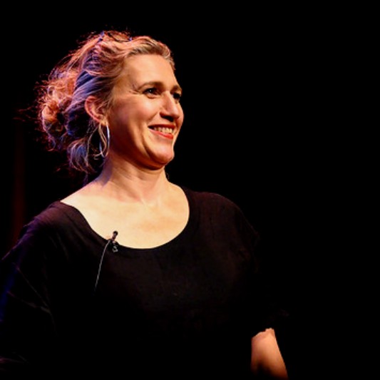 Louise Bloom has short blonde hair tied back. She is smiling and wearing a black top. She is presenting to an audience.